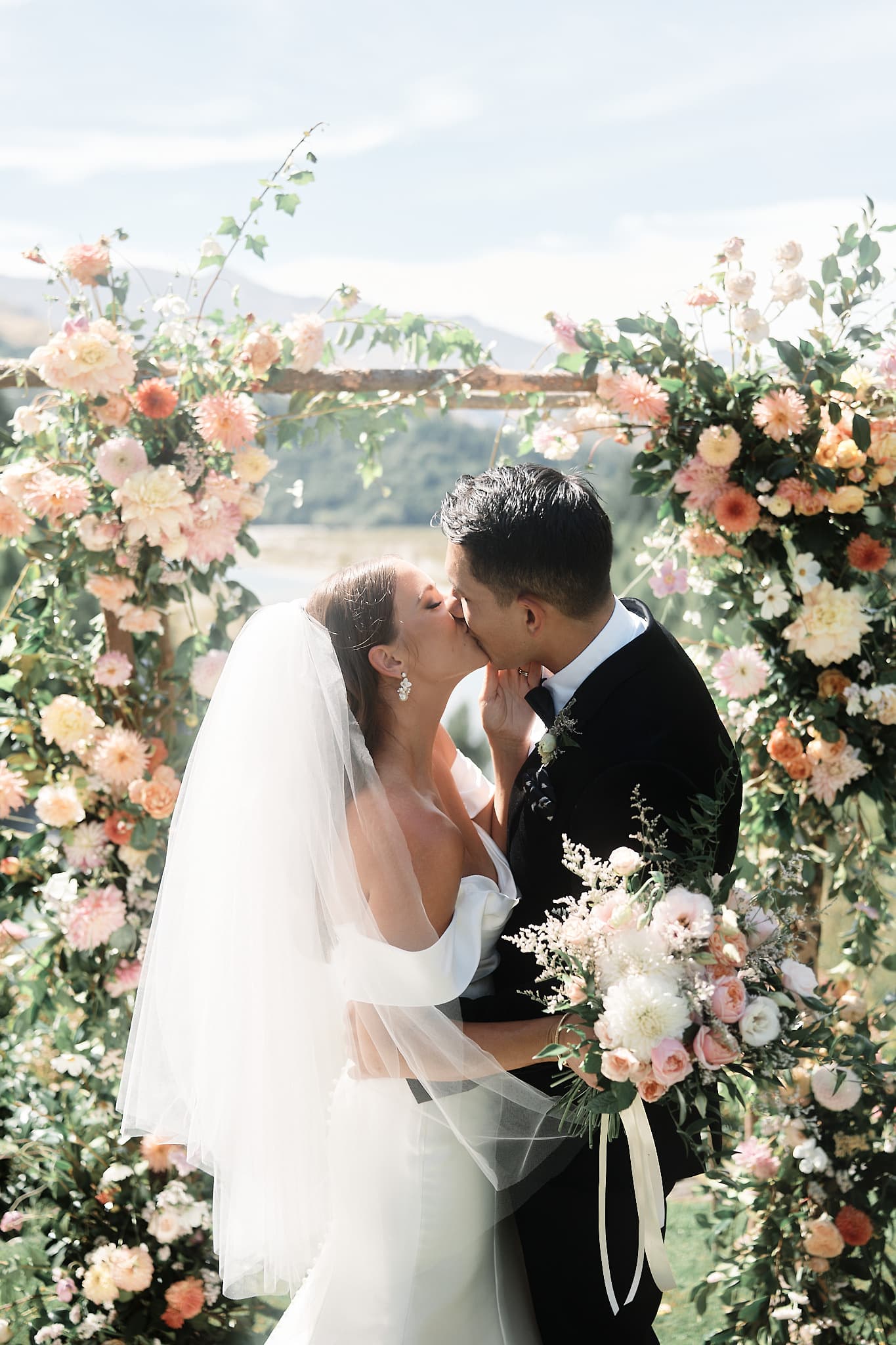 クイーンズタウン　ニュージーランド　ウェディング　前撮り　挙式　結婚式　フォトグラファー | クイーンズタウンのヘリウェディングで、花のアーチの下でキスをする新郎新婦。
